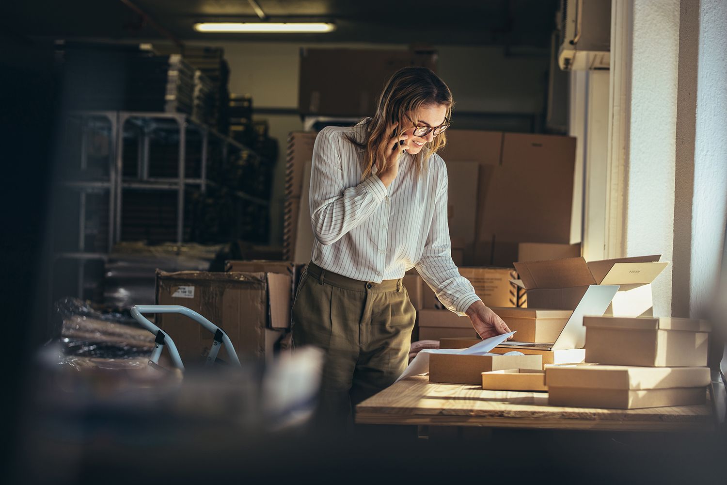 Woman online seller confirming orders from customer on the phone. E-commerce business owner looking at the papers and talking on phone in store warehouse.