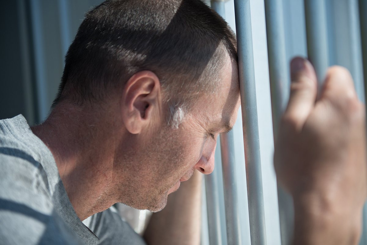 Depressed male inmate holds prison bars in despair