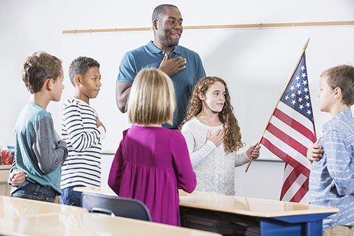 An African American teacher and a multi-ethnic group of elementary school children standing in the classroom saying the pledge of allegiance. A 9 year old Hispanic girl is holding the American flag, hand on heart.