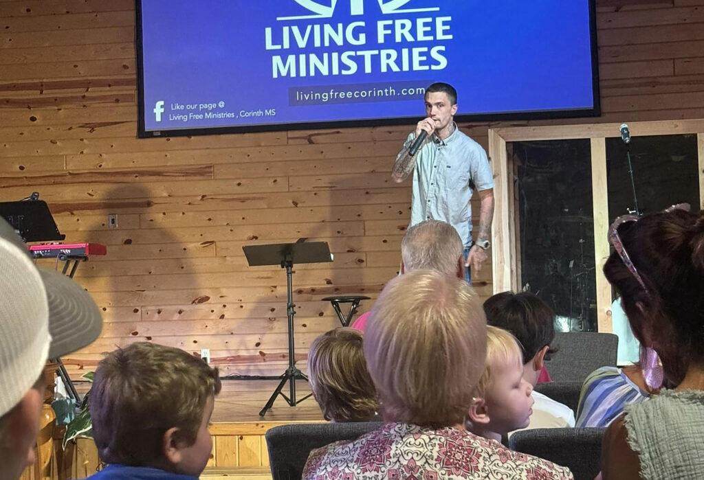 A man speaking on a stage in front of a Living Free Ministries screen, with an audience watching