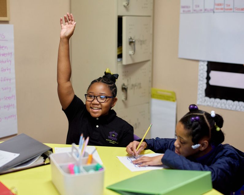 A girl raising her hand while classmates write at a shared table