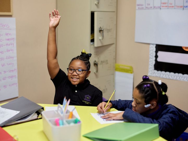 A girl raising her hand while classmates write at a shared table