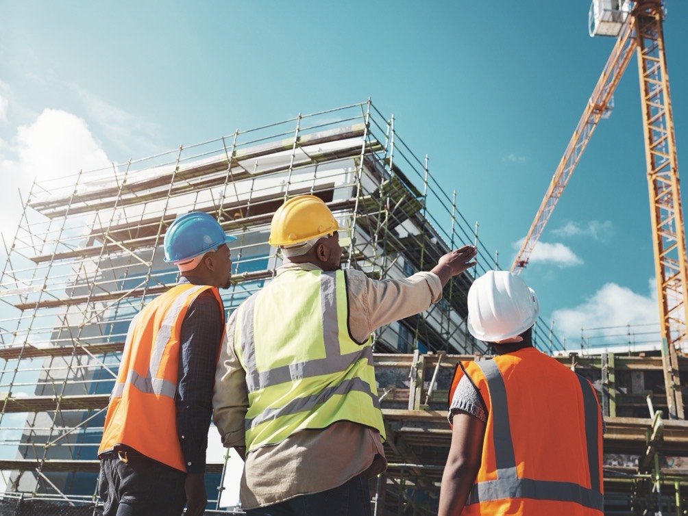 Three construction workers in hard hats and high-visibility vests looking up at a building under scaffolding, one of them pointing toward a crane