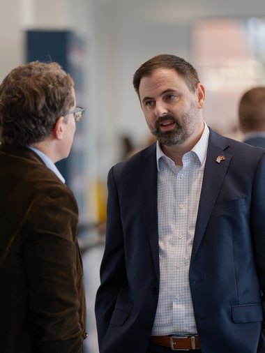 Two people talking at an event, one in a navy suit with a flag lapel pin
