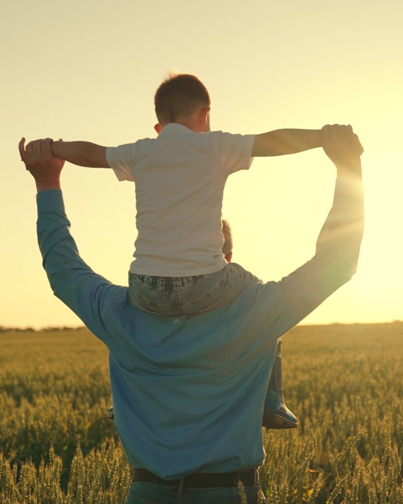 A father carries his son on his shoulders through a field of wheat at sunset, the boy’s arms outstretched