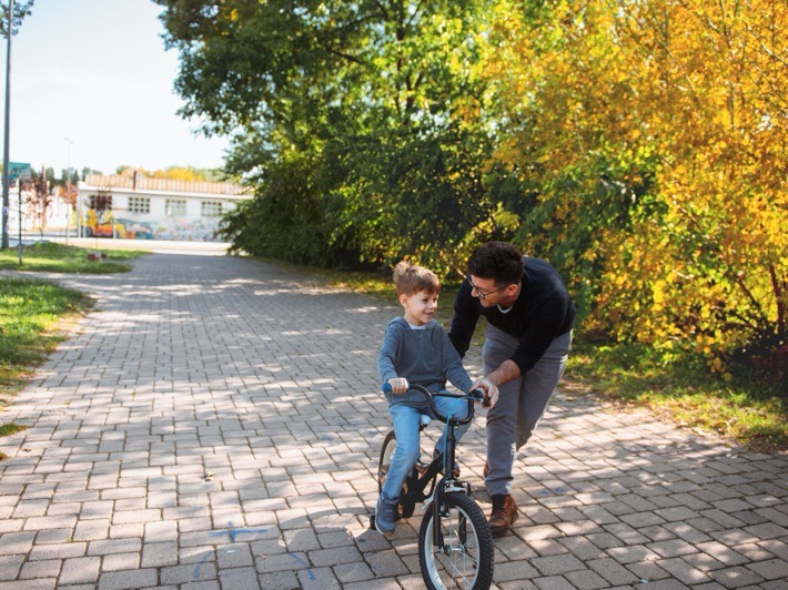 A father steadying his son on a bicycle along a paved park path in autumn