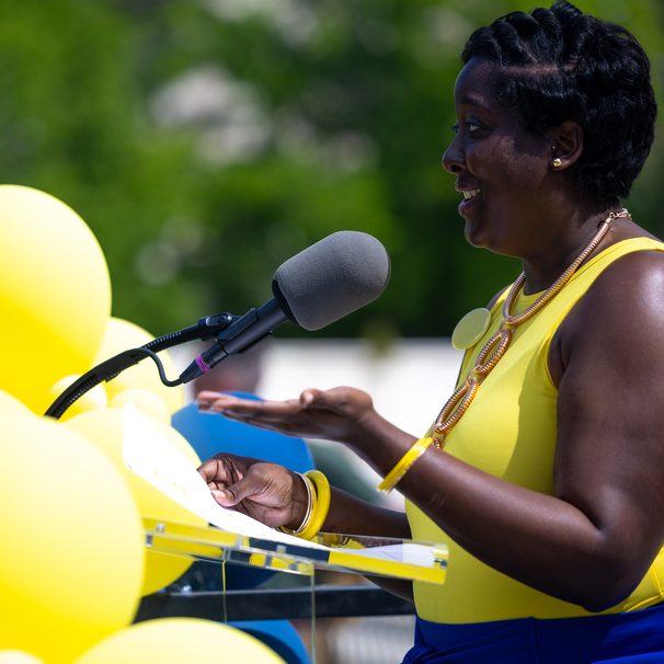 A woman speaking into a microphone at an outdoor event, beside a cluster of yellow balloons